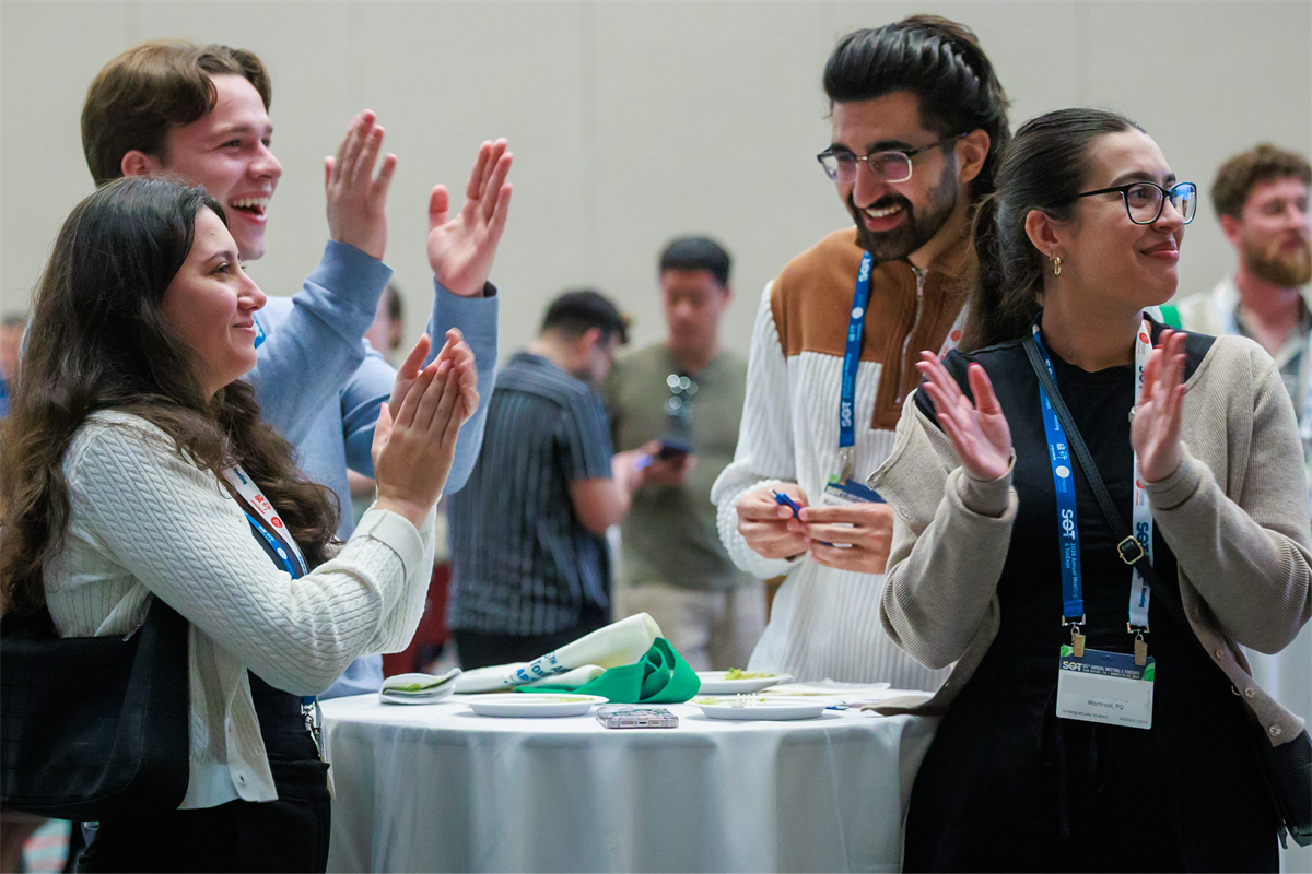Four people standing at a table, smiling and clapping their hands.